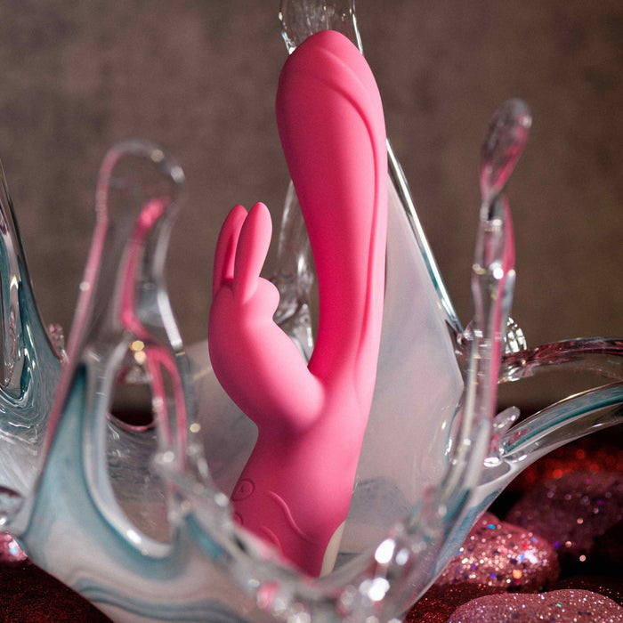 The Bunny Kisses Rechargeable Silicone Rabbit Vibrator stands upright amid a dynamic splash and sparkling pink stones, designed for intense clitoral stimulation against a blurred dark background.
