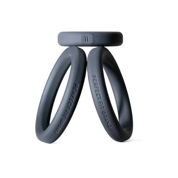 Three black silicone rings from the Perfect Fit Play Zone Cock Ring Kit stand upright and lean against each other, with a third flat ring balanced on top. All three rings have "PERFECT FIT BRAND" labels and are set against a white background.