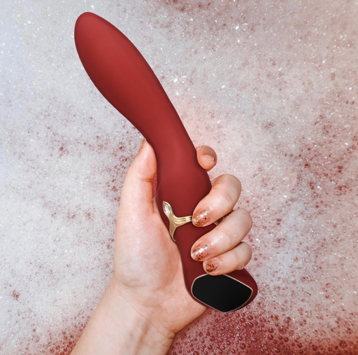 A hand with glittery nail polish holds the maroon Chance Waterproof Touch Screen G-Spot Vibrator, crafted from medical-grade silicone with gold accents, against a bubbly foam background.
