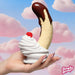 A hand with long, glittery nails holds the Chocolate Dipped Banana Split Silicone Dildo with Suction Cup, standing upright in whipped cream and a cherry, set against a pink cloud sky. The Simply Sweet logo appears in the corner.