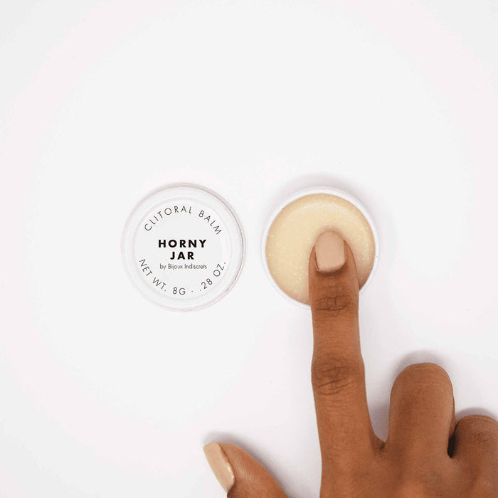 A manicured hand dips a finger into an open Clitherapy Horny Jar Clitoral Balm – Sandalwood Scent, with the lid beside the jar on a white background, highlighting its warming sensation formula.