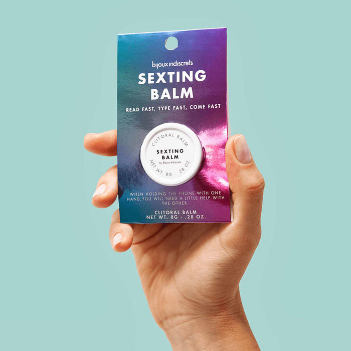 A hand holds a small tin of Clitherapy Sexting Clitoral Balm - Spiced Ginger by Bijoux Indiscrets, presented on a vibrant blue and purple card. The balm is designed to enhance sensitivity during intimate moments, with a light blue background.