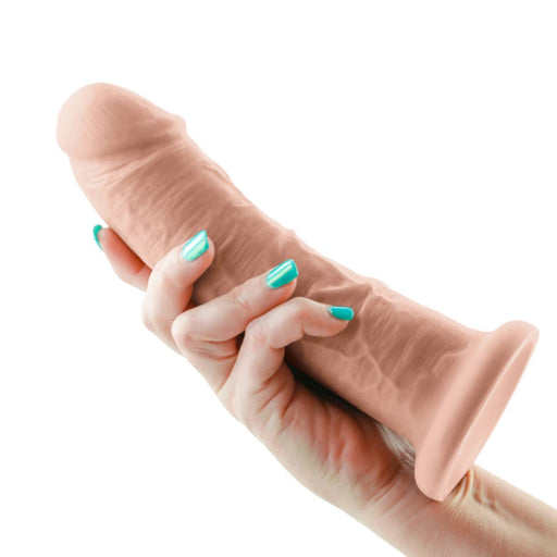 A hand with teal-painted nails holds the Colours Girthy 8 Inch Dual Density Light Tone Silicone Suction Cup Dildo, showing its realistic look and harness compatibility against a white background.