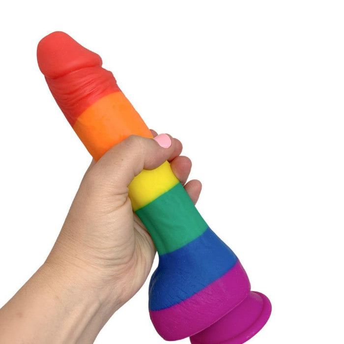 A hand holds the Colours Pride Rainbow Long Thick Silicone 9.5 Inch Dildo, featuring a suction cup base, against a plain white background.