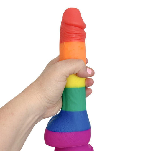 A hand holds the Colours Pride Rainbow Long Thick Silicone 9.5 Inch Dildo, shaped like a phallus, against a white background.