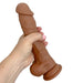 A hand holds the Colours Realistic Long 9.5 inch Medium Tone Silicone Dildo, featuring a girthy design and suction cup base for strap-on harness compatibility, set against a white background.