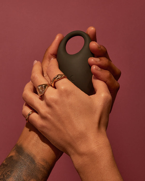Two hands with noticeable jewelry holding a modern, sleek black Pepper Connect Vibrating Cock Ring against a warm-toned backdrop.