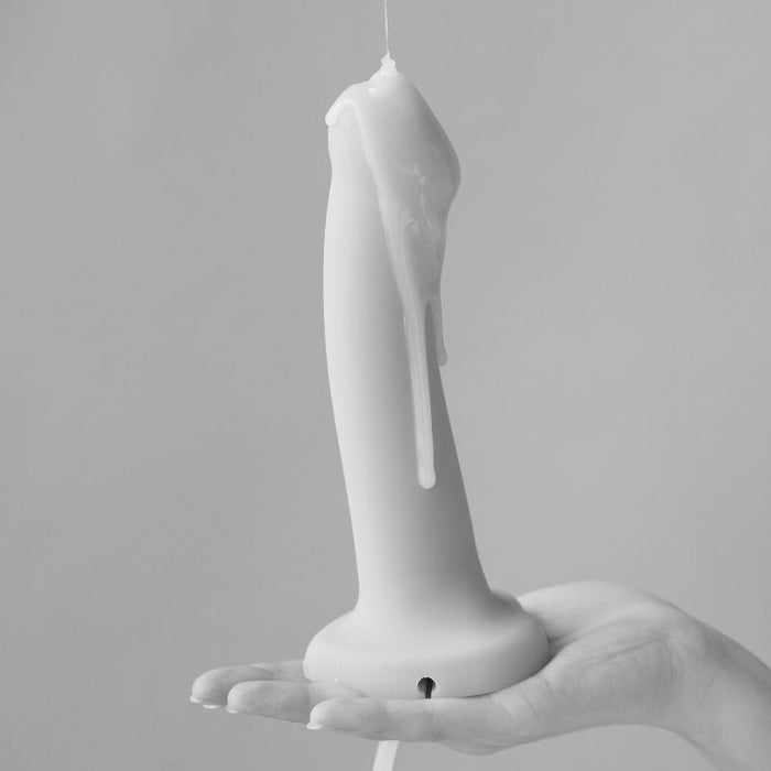 A hand holds an upright bottle of Creamy Glide Milky Semen-Style Cum Lubricant (8.5 fl oz), with the smooth, creamy liquid dripping down its side. The background is plain and softly blurred in this black and white image.