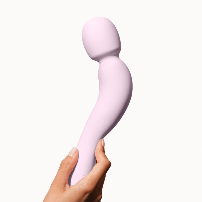 A hand holds the Dame Com Ergonomic Waterproof Wand Vibrator, a light pink device with a flexible neck and rounded head for deep rumbly clitoral pleasure, set against a plain white background.