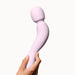 A hand holds the Dame Com Ergonomic Waterproof Wand Vibrator, a light pink device with a flexible neck and rounded head for deep rumbly clitoral pleasure, set against a plain white background.