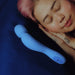 A person with pink hair sleeps peacefully on a dark blue pillow, hands under their face. Beside them lies the Dame Com Ergonomic Waterproof Wand Vibrator with Flexible Neck, known for deep rumbly clitoral pleasure.