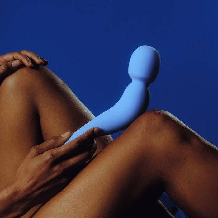 Someone holds the Dame Com Ergonomic Waterproof Wand Vibrator with Flexible Neck—a curved blue silicone toy—against their bare thigh, legs crossed, set against a solid blue background.