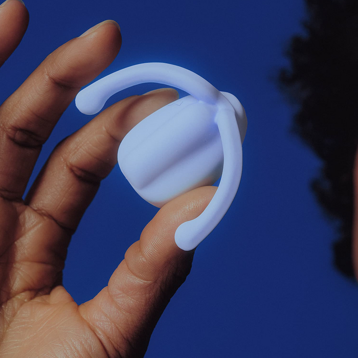 A hand holds the Dame Eva Hands-Free Silicone Clitoral Vibrator in Ice Blue, a small wearable device with two flexible, curved arms, shown against a dark blue background.