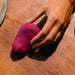 A hand reaches for the Dame Pom Hand Held Flexible Vibrator in Plum, a small, purple waterproof toy with water droplets on it, resting on a wooden surface.