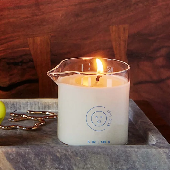 A lit Melt Together Massage Oil Candle by Dame (Green Tea, Fig & Cedarwood) in a clear beaker with blue markings and the Wild Lust logo sits on a gray tray, with gold décor and a green ball partially visible beside it.