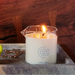 A lit Melt Together Massage Oil Candle by Dame (Green Tea, Fig & Cedarwood) in a clear beaker with blue markings and the Wild Lust logo sits on a gray tray, with gold décor and a green ball partially visible beside it.