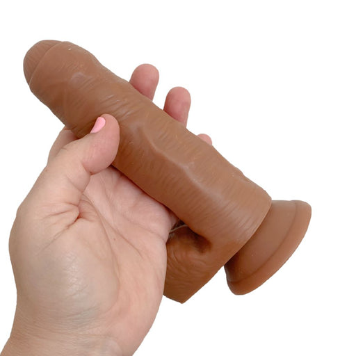 A hand holds the Derek the Bartender Lifelike Uncut 7 Inch Medium Tone Dildo, featuring realistic detail and a flared suction cup base, against a white background.