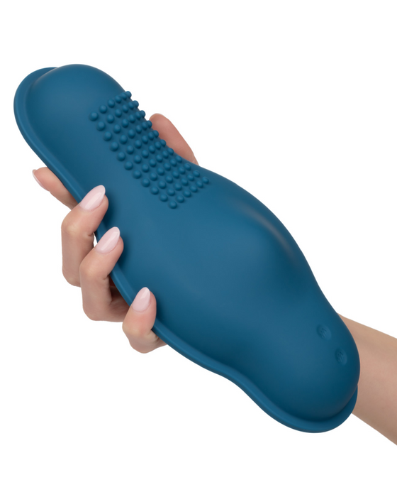 A hand holding a blue silicone pet grooming brush with textured bristles on the surface. The brush, much like the CalExotics Dual Rider Remote Control Bump & Grind Humping Vibrator, features an ergonomic design for easy handling.