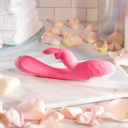 The Bunny Kisses Rechargeable Silicone Rabbit Vibrator, featuring a pink curved silicone design and rabbit-style extension for clitoral and G-spot stimulation, rests on a marble surface with rose petals and folded white towels nearby.