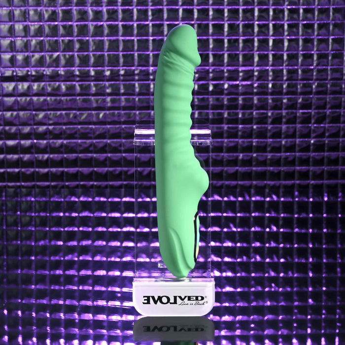 The Full Throttle Warming Waterproof Silicone Vibrator, in green with a ribbed shaft, sits on a clear stand labeled "EVOLVED" against a reflective purple tiled background under dramatic lighting.