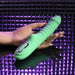 A hand holds the Full Throttle Warming Waterproof Silicone Vibrator, featuring control buttons and a ribbed green silicone shaft, set against a shiny purple textured background.