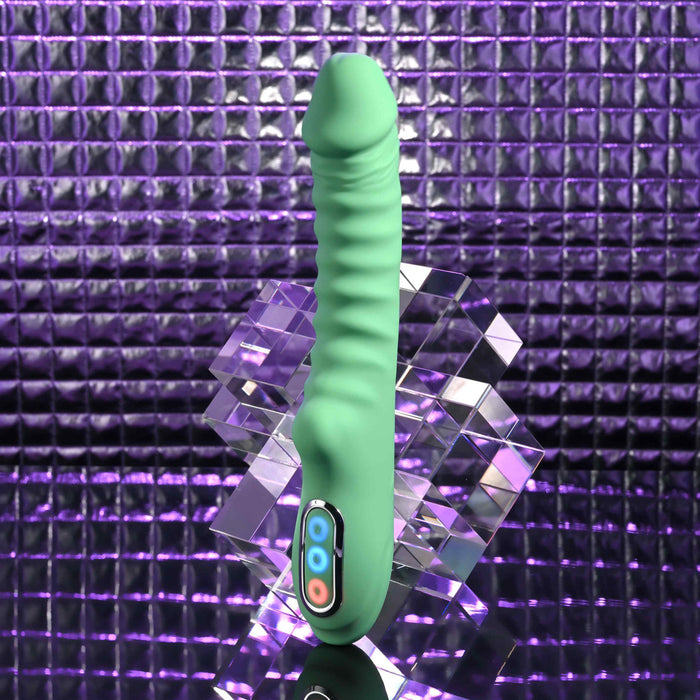 The Full Throttle Warming Waterproof Silicone Vibrator, in green with a ribbed shaft and control buttons, is shown upright on a reflective geometric surface against a purple textured background.