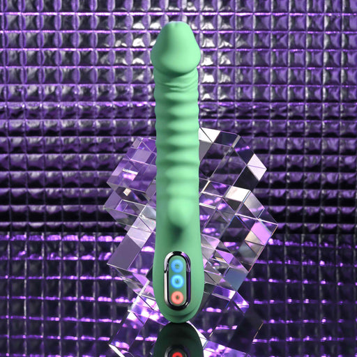 The Full Throttle Warming Waterproof Silicone Vibrator, featuring green ribbed silicone and a control panel at the base, stands upright by a geometric acrylic block against a purple, metallic-textured background.