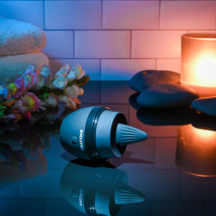 The Tongue Tied Licking Tongue Rim Job Vibrator sits on a glossy surface near stacked black stones, a lit candle, soft towels, and flowers, creating a soothing spa-like setting with gentle, colorful lighting.