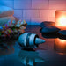 The Tongue Tied Licking Tongue Rim Job Vibrator sits on a glossy surface near stacked black stones, a lit candle, soft towels, and flowers, creating a soothing spa-like setting with gentle, colorful lighting.