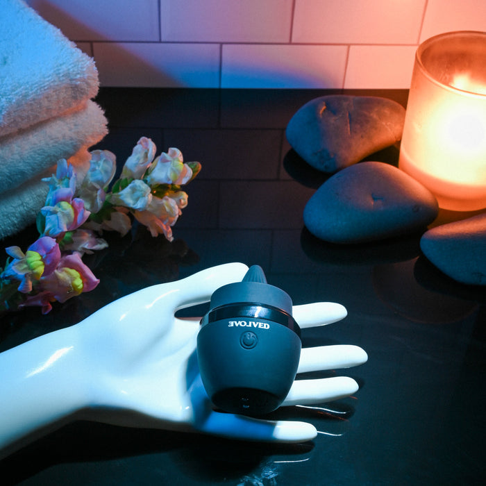 The Tongue Tied Licking Tongue Rim Job Vibrator in black sits on a white mannequin hand amid smooth stones, pink flowers, folded towels, and a glowing candle on a glossy surface in a spa-like setting.