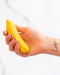 A hand holding a Banana Emojibator Vibrator with a tattoo that reads "love" on the wrist. The background is a blurred white and gray pattern.