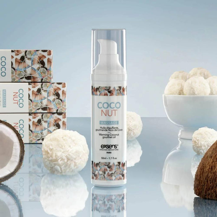 A bottle of Exsens Coconut Flavored Warming Massage Oil 50ml stands on a reflective surface amid coconut halves, white coconut balls, and product boxes against a light blue background—crafted with vegan ingredients for pure indulgence.