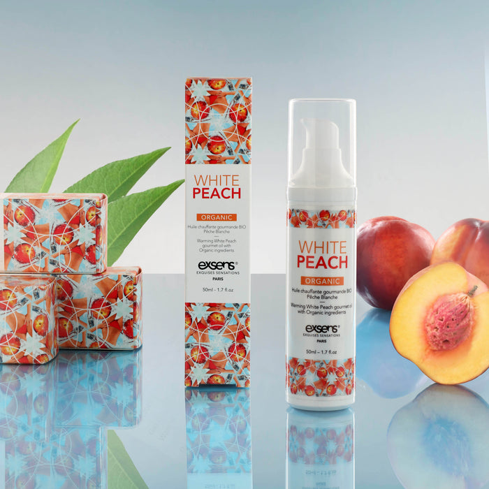 A bottle and box of Exsens Organic White Peach Flavored Warming Massage Oil 50ml are shown with fresh peaches, a sliced peach, aloe vera leaves, and decorative soap bars against a light blue background.