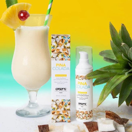 A pina colada cocktail garnished with pineapple and cherry sits beside a box and bottle of Exsens Pina Colada Flavored Warming Massage Oil 50ml on a yellow and teal background, with coconut pieces and a pineapple in the foreground.