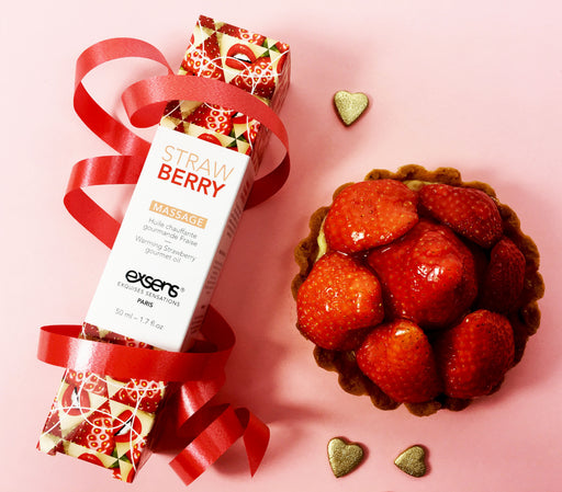 A box of Exsens Strawberry Flavored Warming Massage Oil 50ml, wrapped with a red ribbon, is displayed beside a strawberry tart and two gold heart-shaped decorations on a pink background.