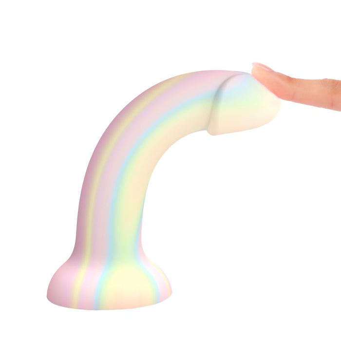 A person's finger gently touches the tip of the Fantasia Smooth 7 inch Glow in the Dark Silicone Dildo with Suction Cup, which features a pastel rainbow design and curved shape, set against a white background.