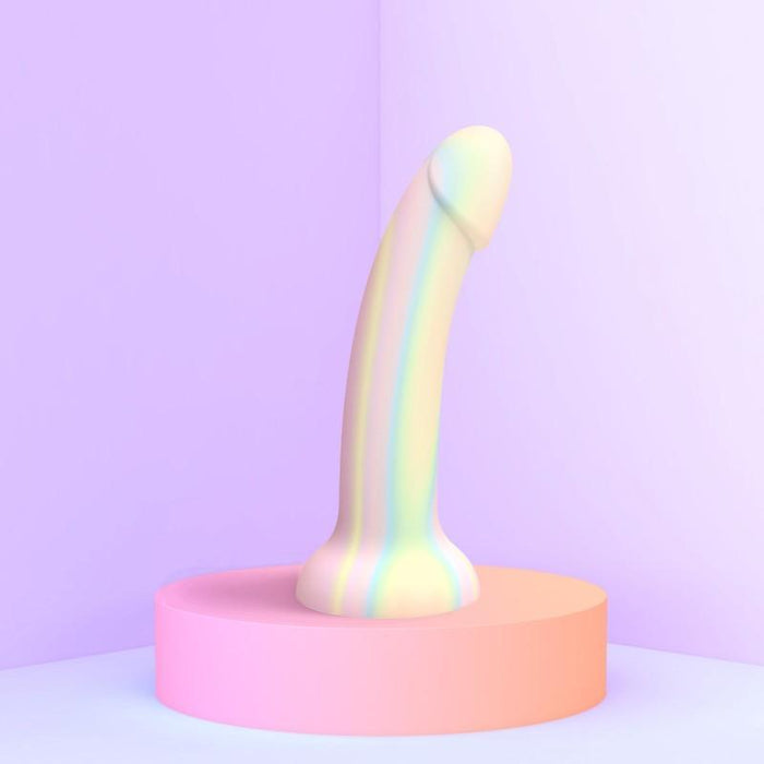 The Fantasia Smooth 7 inch Glow in the Dark Silicone Dildo with Suction Cup, featuring pastel rainbow stripes, stands upright on a pink platform against a soft purple backdrop in a bright, minimalistic scene.