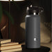 A black matte insulated **Fuck Flask Pussy Stroker Disguised as a Water Bottle - Vanilla** by **Pipedream Products** with a lid and handle sits discreetly on a black table next to a small stack of books. A nearby lamp casts a warm light over the scene, creating a cozy ambiance. In the background, a pillow and part of a sofa are visible.