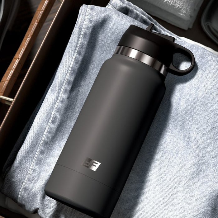A black Fuck Flask Pussy Stroker Disguised as a Water Bottle - Vanilla by Pipedream Products with a loop cap is placed on top of a pair of folded light blue jeans in a wooden drawer. Nearby are neatly arranged towels and other items, all meticulously organized. The water bottle, bearing an "EE" logo on its side, remains discreet within the tidy setting.