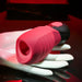 The Evolved Novelties Body Kisses Red Suction Masturbator and Vibrator, featuring a pink and black design, rests on a white mannequin hand. The item has a soft, open-ended structure and is prominently displayed against a dark background with red lighting.
