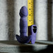 The Ring It Remote Control Rimming Vibrating Prostate Plug in purple stands upright beside a yellow measuring tape and its remote control, all set against a textured gray background.