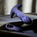 The Ring It Remote Control Rimming Vibrating Prostate Plug, a purple silicone massager with a curved, ridged design and wide base, rests on stone beside its black remote. Soft light and shadows highlight this sleek prostate toy.