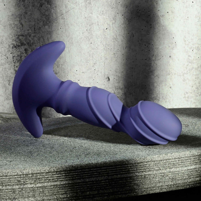The Ring It Remote Control Rimming Vibrating Prostate Plug, a purple silicone massager with a curved handle and ridged shaft, sits on a textured gray surface against a concrete wall background.