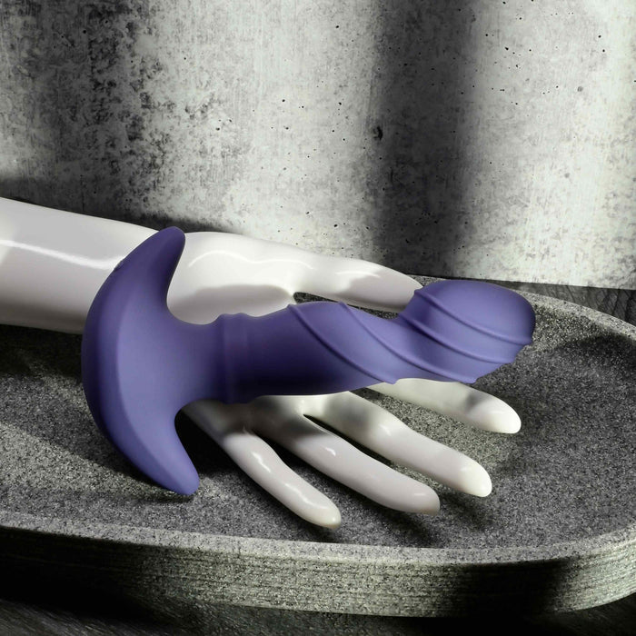 The Ring It Remote Control Rimming Vibrating Prostate Plug, made of purple silicone with a curved, textured design, is displayed on a white mannequin hand in a gray stone tray against a concrete wall and curtain.