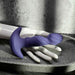 The Ring It Remote Control Rimming Vibrating Prostate Plug, made of purple silicone with a curved, textured design, is displayed on a white mannequin hand in a gray stone tray against a concrete wall and curtain.