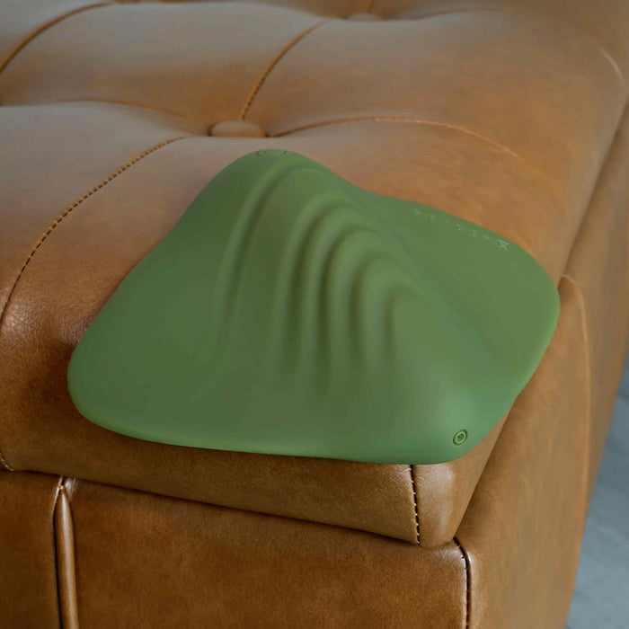 A green, mountain-shaped Gender X Pleasure Pad Rechargeable Grinding Pad & Stroker with wavy ridges sits on the corner of a tan, tufted leather ottoman.
