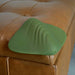 A green, mountain-shaped Gender X Pleasure Pad Rechargeable Grinding Pad & Stroker with wavy ridges sits on the corner of a tan, tufted leather ottoman.