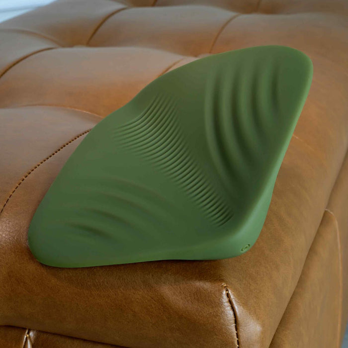 A green Gender X Pleasure Pad Rechargeable Grinding Pad & Stroker with textured ridges sits on a tan leather tufted ottoman.