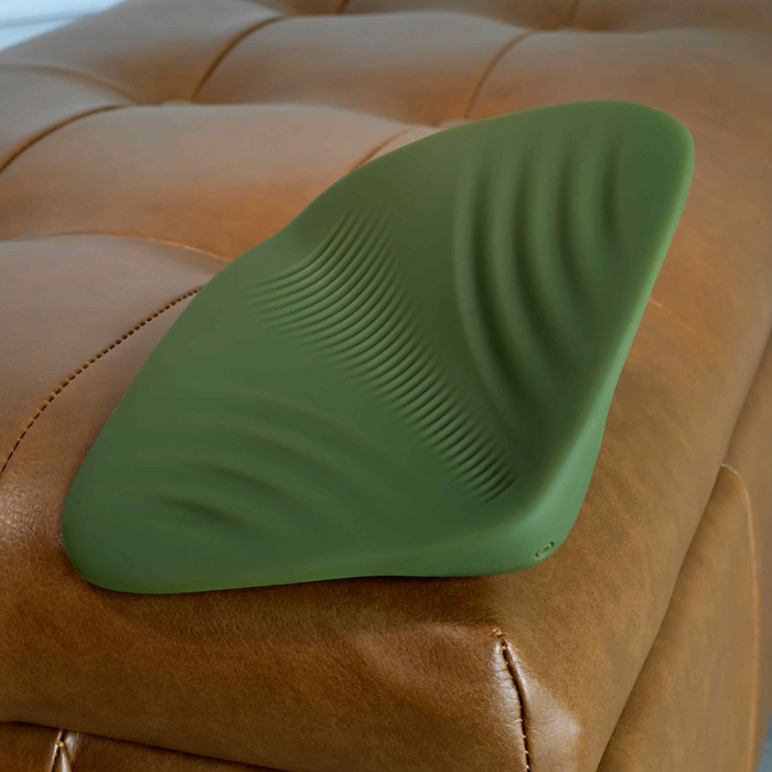 A green Gender X Pleasure Pad Rechargeable Grinding Pad & Stroker with textured ridges sits on a tan leather tufted ottoman.