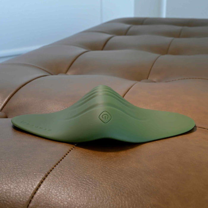 The Gender X Pleasure Pad Rechargeable Grinding Pad & Stroker, featuring a green, curved silicone design with smooth ridges and a power button symbol, rests on a tan tufted leather surface. The device showcases a modern minimalist look.